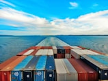 A vast ocean with a cargo ship moving forward, leaving a trail in the water. The deck is packed with organized shipping containers in various colors, including red, blue, and white. The sky is partly cloudy with a vibrant blue hue, creating a serene and expansive horizon.