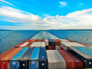 A vast ocean with a cargo ship moving forward, leaving a trail in the water. The deck is packed with organized shipping containers in various colors, including red, blue, and white. The sky is partly cloudy with a vibrant blue hue, creating a serene and expansive horizon.