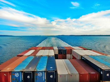A vast ocean with a cargo ship moving forward, leaving a trail in the water. The deck is packed with organized shipping containers in various colors, including red, blue, and white. The sky is partly cloudy with a vibrant blue hue, creating a serene and expansive horizon.