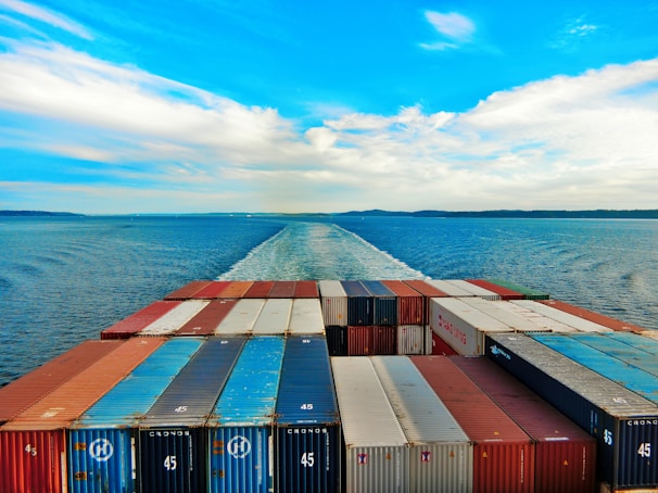A vast ocean with a cargo ship moving forward, leaving a trail in the water. The deck is packed with organized shipping containers in various colors, including red, blue, and white. The sky is partly cloudy with a vibrant blue hue, creating a serene and expansive horizon.
