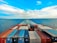 A vast ocean with a cargo ship moving forward, leaving a trail in the water. The deck is packed with organized shipping containers in various colors, including red, blue, and white. The sky is partly cloudy with a vibrant blue hue, creating a serene and expansive horizon.