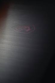 A dark surface with a subtle red emblem of a dragon inside an angular shield shape, likely indicating a brand logo. The texture is smooth with a slightly brushed metallic finish, giving a sleek and modern appearance.