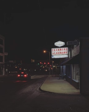 A nighttime urban street scene featuring a brightly lit sign for a dry cleaners and laundry service. The glow from the sign illuminates the adjacent sidewalk and part of the building. Traffic lights and car headlights are visible in the background, providing a sense of depth and perspective.