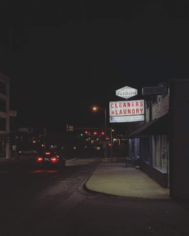 A nighttime urban street scene featuring a brightly lit sign for a dry cleaners and laundry service. The glow from the sign illuminates the adjacent sidewalk and part of the building. Traffic lights and car headlights are visible in the background, providing a sense of depth and perspective.