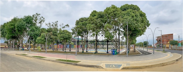 A spacious urban park area with tall, lush green trees lined along a paved walkway. There's an enclosed sports court in the center surrounded by a fence. Modern streetlights and seating areas are visible. Buildings and houses are located in the background across a road, adding to the neighborhood setting.