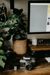 A vintage camera rests on a wooden desk surrounded by photography equipment, including a film roll and lenses. A large leafy plant in a woven basket adds a touch of nature to the scene. A computer monitor occupies part of the desk, and the overall setting appears to be a workspace combining technology and nature.