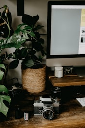 A vintage camera rests on a wooden desk surrounded by photography equipment, including a film roll and lenses. A large leafy plant in a woven basket adds a touch of nature to the scene. A computer monitor occupies part of the desk, and the overall setting appears to be a workspace combining technology and nature.