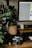 A vintage camera rests on a wooden desk surrounded by photography equipment, including a film roll and lenses. A large leafy plant in a woven basket adds a touch of nature to the scene. A computer monitor occupies part of the desk, and the overall setting appears to be a workspace combining technology and nature.