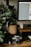 A vintage camera rests on a wooden desk surrounded by photography equipment, including a film roll and lenses. A large leafy plant in a woven basket adds a touch of nature to the scene. A computer monitor occupies part of the desk, and the overall setting appears to be a workspace combining technology and nature.