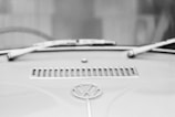 A monochrome close-up of a vintage car hood, featuring the Volkswagen logo prominently in the center. The windshield wipers are visible along the top, and there's a rectangular vent just above the emblem.