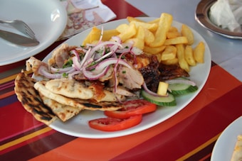 A plate containing grilled meat topped with chopped red onions and garnished with parsley, accompanied by slices of cucumber and tomato. Beside the meat, there are crispy golden french fries and pieces of flatbread, all served on a white plate. The setting includes a colorful striped tablecloth with a fork and spoon placed on a plate nearby.