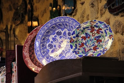 Intricately designed decorative plates with vibrant patterns are displayed in a row against a textured stone wall. The foreground features a prominent blue plate with floral motifs, flanked by a multicolored plate on the right and a maroon plate on the left.