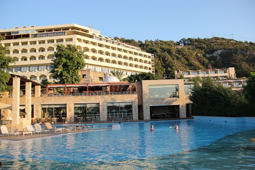 A large hotel is situated next to a swimming pool with lounge chairs arranged along its edge. The hotel features multiple stories with rows of balconies. Lush greenery and trees surround the area. The swimming pool is clear and inviting, with a couple of people swimming and playing with a ball. The sun casts warm, golden light over the scene, indicating either morning or late afternoon.