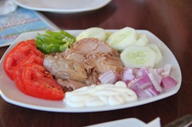 A plate of food featuring neatly arranged slices of fresh tomatoes, cucumber, and red onion alongside chunks of canned tuna. A dollop of mayonnaise is placed on the side, and a few slices of green bell pepper add color to the dish.