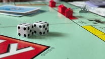 A board game in progress featuring Monopoly with two dice showing different numbers. Red plastic houses are placed on the board, and stacks of cards are visible. The board is vibrant with several distinct colors and clear outlines of property names.