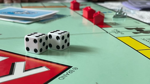 A board game in progress featuring Monopoly with two dice showing different numbers. Red plastic houses are placed on the board, and stacks of cards are visible. The board is vibrant with several distinct colors and clear outlines of property names.