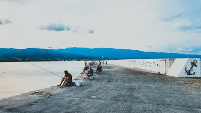 Several people are fishing along a concrete pier or breakwater that extends out into a body of water. The scene is set against a backdrop of distant mountains under a lightly clouded sky. Graffiti artwork is visible on the side of the pier.