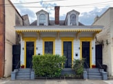 A charming duplex-style house with two front doors, painted in white with yellow trim. It features decorative elements under the eaves and two dormer windows on the roof. Two small staircases lead to each door, flanked by potted plants and a central bush. The house is situated between two larger buildings and has a brick chimney in the center.