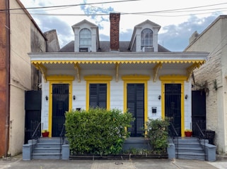 A charming duplex-style house with two front doors, painted in white with yellow trim. It features decorative elements under the eaves and two dormer windows on the roof. Two small staircases lead to each door, flanked by potted plants and a central bush. The house is situated between two larger buildings and has a brick chimney in the center.