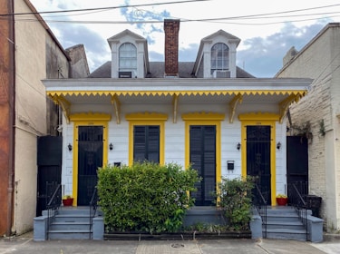A charming duplex-style house with two front doors, painted in white with yellow trim. It features decorative elements under the eaves and two dormer windows on the roof. Two small staircases lead to each door, flanked by potted plants and a central bush. The house is situated between two larger buildings and has a brick chimney in the center.