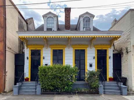 A charming duplex-style house with two front doors, painted in white with yellow trim. It features decorative elements under the eaves and two dormer windows on the roof. Two small staircases lead to each door, flanked by potted plants and a central bush. The house is situated between two larger buildings and has a brick chimney in the center.