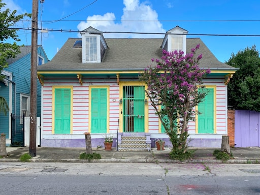 A vibrant, colorful house with teal shutters and doors, yellow trim, and a purple base. The exterior is striped with alternating light pink and white horizontal lines. Two small dormer windows protrude from the roof, and a flowering tree stands to the side. Potted plants and a small set of tiled steps lead up to the entrance.