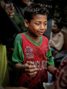 A young boy wears a red and green sports jersey with the word 'Bangladesh' on it. The jersey features a logo for the ICC World Twenty20 event in 2016. He has a focused and engaged expression on his face, with his hands slightly raised.