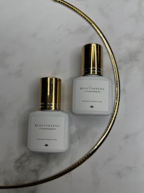 Two white bottles with gold caps are placed on a marble surface. The bottles have the label 'BEAUTYRAENA' and mention a drying time of 1-2 seconds. A thin gold curved line is present in the background.