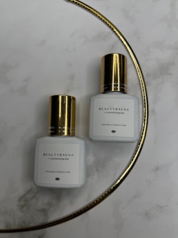Two white bottles with gold caps are placed on a marble surface. The bottles have the label 'BEAUTYRAENA' and mention a drying time of 1-2 seconds. A thin gold curved line is present in the background.