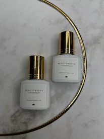 Two white bottles with gold caps are placed on a marble surface. The bottles have the label 'BEAUTYRAENA' and mention a drying time of 1-2 seconds. A thin gold curved line is present in the background.