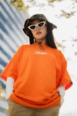 A person wearing an orange T-shirt with white writing, layered over a long-sleeve white shirt. They have long dark hair and are accessorized with white cat-eye sunglasses and a camouflage bucket hat. The background includes a modern building with blue tinted windows and some tree branches.