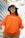 A person wearing an orange T-shirt with white writing, layered over a long-sleeve white shirt. They have long dark hair and are accessorized with white cat-eye sunglasses and a camouflage bucket hat. The background includes a modern building with blue tinted windows and some tree branches.