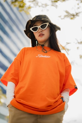 A person wearing an orange T-shirt with white writing, layered over a long-sleeve white shirt. They have long dark hair and are accessorized with white cat-eye sunglasses and a camouflage bucket hat. The background includes a modern building with blue tinted windows and some tree branches.