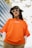 A person wearing an orange T-shirt with white writing, layered over a long-sleeve white shirt. They have long dark hair and are accessorized with white cat-eye sunglasses and a camouflage bucket hat. The background includes a modern building with blue tinted windows and some tree branches.
