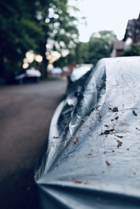 A close-up of a covered car parked on a residential street, with trees and houses visible in the blurred background. The car cover appears worn and has dirt on it, suggesting it has been there for some time.