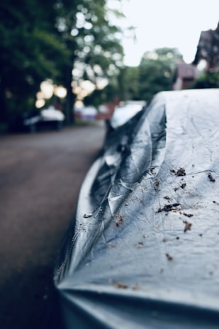 A close-up of a covered car parked on a residential street, with trees and houses visible in the blurred background. The car cover appears worn and has dirt on it, suggesting it has been there for some time.