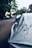 A close-up of a covered car parked on a residential street, with trees and houses visible in the blurred background. The car cover appears worn and has dirt on it, suggesting it has been there for some time.