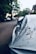 A close-up of a covered car parked on a residential street, with trees and houses visible in the blurred background. The car cover appears worn and has dirt on it, suggesting it has been there for some time.