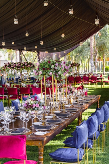 A long, elegantly set dining table is placed under a tented canopy. The table is adorned with multiple wine glasses, plates, and floral centerpieces featuring pink and red flowers. Tall candlesticks add height to the table setting. Plush chairs with blue and pink velvet cushions surround the table. Hanging glass lanterns are lit, adding a warm ambiance. The background features palm trees and additional decorated tables.