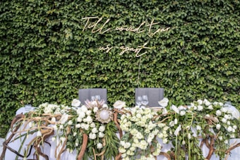 A lush green wall covered with dense foliage serves as the backdrop. In front, there is a table adorned with an abundant arrangement of white roses, lilies, and intertwining branches. A neon sign with elegant cursive writing is mounted on the wall, exuding a warm glow. Wine glasses are set on the table, hinting at a formal or celebratory event.
