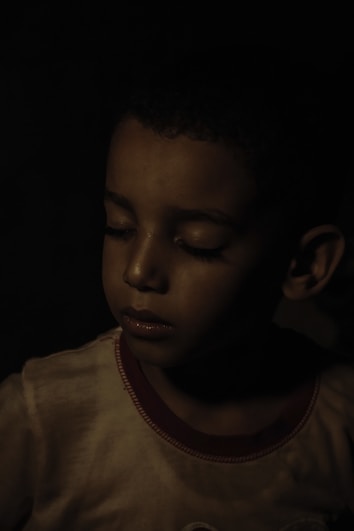 A young child with eyes closed, appearing contemplative or thoughtful, is softly illuminated in a dimly lit environment. The lighting is low-key, creating deep shadows that enhance the mood of solitude or introspection.