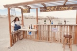 A beachside bar setup with a woman standing beside it. The bar is made of wooden planks, and various drink bottles are displayed on the counter. Signs indicate 'OPEN BAR' and 'SKI INN II'. A stool is positioned in front of the bar. Sand and distant structures are visible in the background.