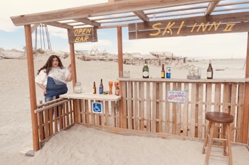 A beachside bar setup with a woman standing beside it. The bar is made of wooden planks, and various drink bottles are displayed on the counter. Signs indicate 'OPEN BAR' and 'SKI INN II'. A stool is positioned in front of the bar. Sand and distant structures are visible in the background.