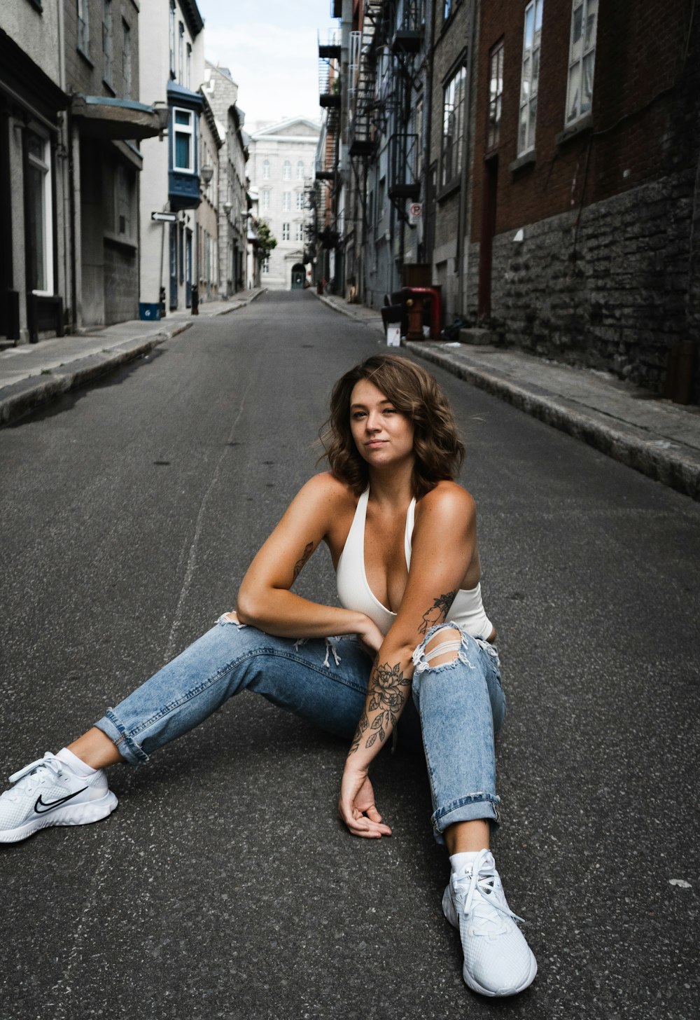 woman in white tank top and blue denim jeans sitting on sidewalk during daytime