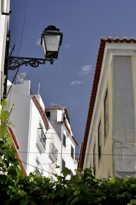 The image features a view of an architectural scene with buildings displaying red-tiled roofs and white walls. A vintage-style street lamp is prominently situated in the foreground on the left. Green foliage is visible at the base, while a clear blue sky forms the background.