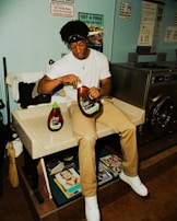 A person sits on a counter in a laundromat, holding a large bottle of fabric softener. Laundry supplies and magazines are visible nearby, along with various signs on the wall, including one offering a free COVID-19 test.