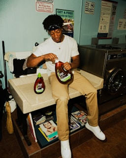 A person sits on a counter in a laundromat, holding a large bottle of fabric softener. Laundry supplies and magazines are visible nearby, along with various signs on the wall, including one offering a free COVID-19 test.