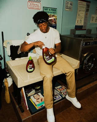 A person sits on a counter in a laundromat, holding a large bottle of fabric softener. Laundry supplies and magazines are visible nearby, along with various signs on the wall, including one offering a free COVID-19 test.