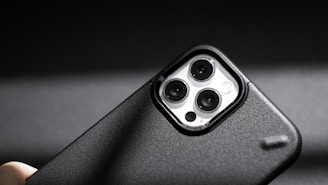 A smartphone camera module with three lenses is prominently featured. The phone case is black with a textured surface, and light reflects off the lens surfaces. The image is sharply focused, with a dark, blurred background that enhances the subject's detail.