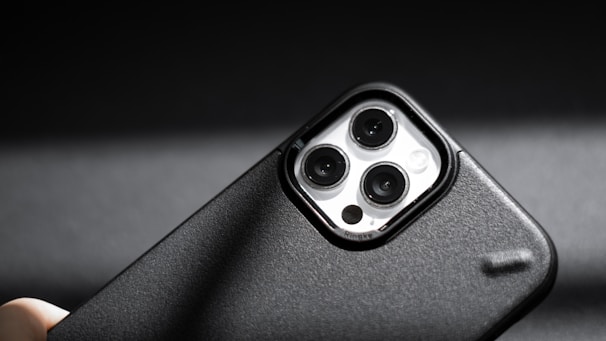 A smartphone camera module with three lenses is prominently featured. The phone case is black with a textured surface, and light reflects off the lens surfaces. The image is sharply focused, with a dark, blurred background that enhances the subject's detail.